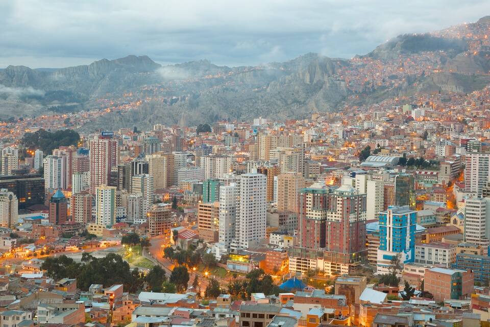 Paisagem urbana com um conjunto denso de edifícios altos, um edifício azul de destaque e montanhas ao fundo.