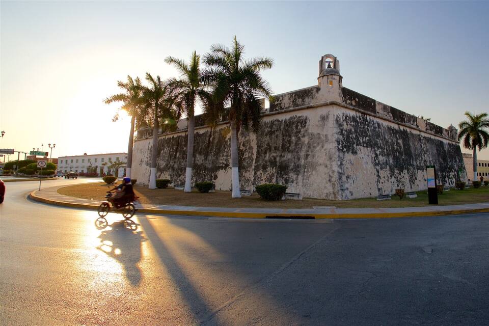 Uma fortaleza histórica com uma torre de sino, palmeiras e uma rotatória com placa de limite de velocidade.