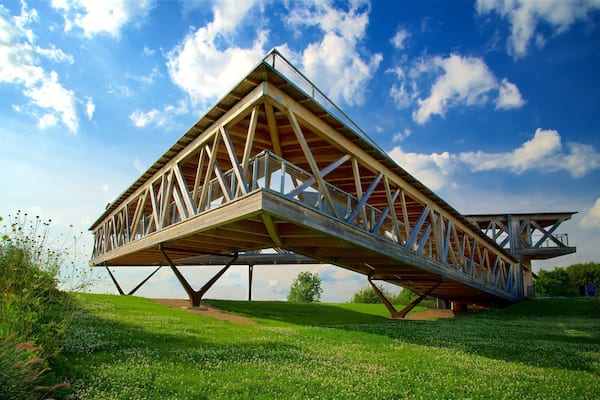 A large wooden bridge with a triangular truss design, supported by a central pillar, spanning a grassy area with wildflowers and trees under a partly cloudy sky.