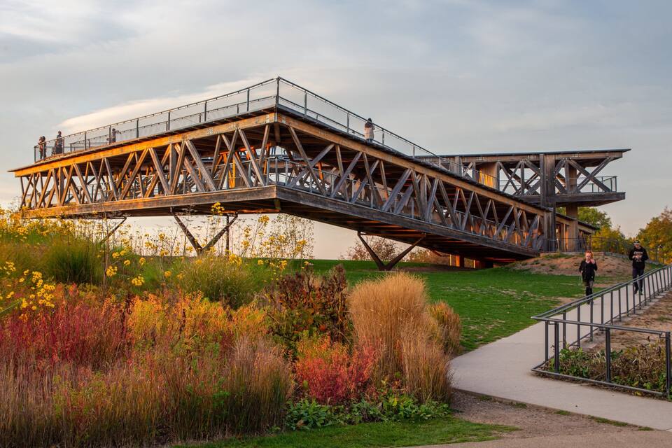 Un pont piétonnier enjambant une rivière, avec une zone herbeuse et des fleurs sauvages en contrebas.