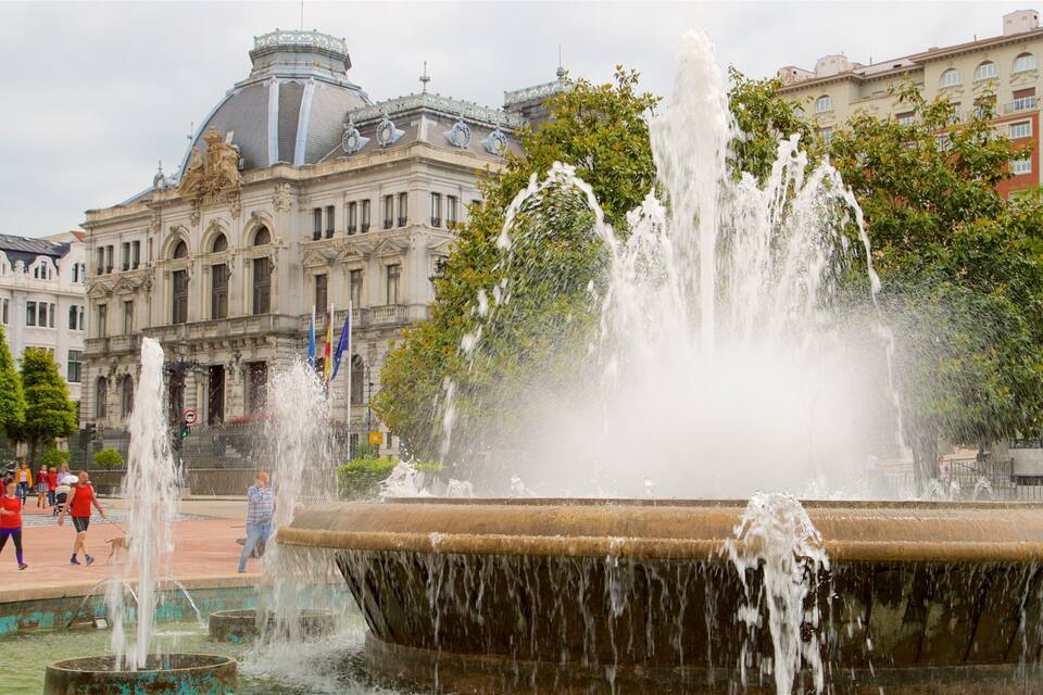 Een grote fontein met waterstralen op een openbaar plein, omringd door historische gebouwen en bomen.