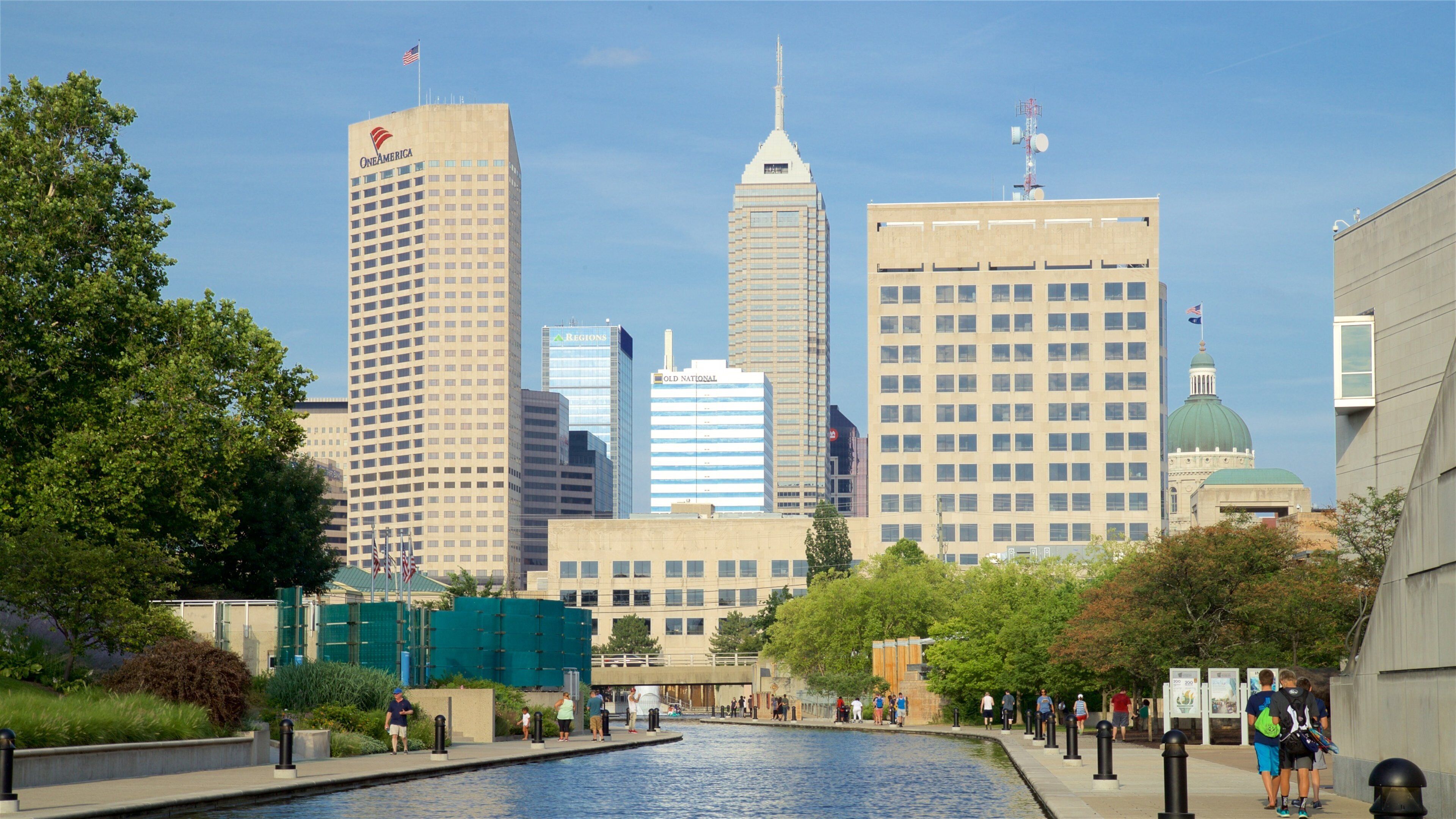 A cityscape with tall buildings, including OneAmerica and ARLINGTONS, and a body of water with people walking along the promenade.