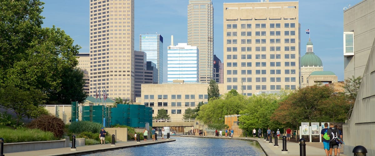 A cityscape with tall buildings, including OneAmerica and ARLINGTONS, and a body of water with people walking along the promenade.