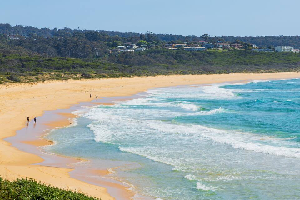 A sandy beach with waves, a forested hill, and houses in the background.