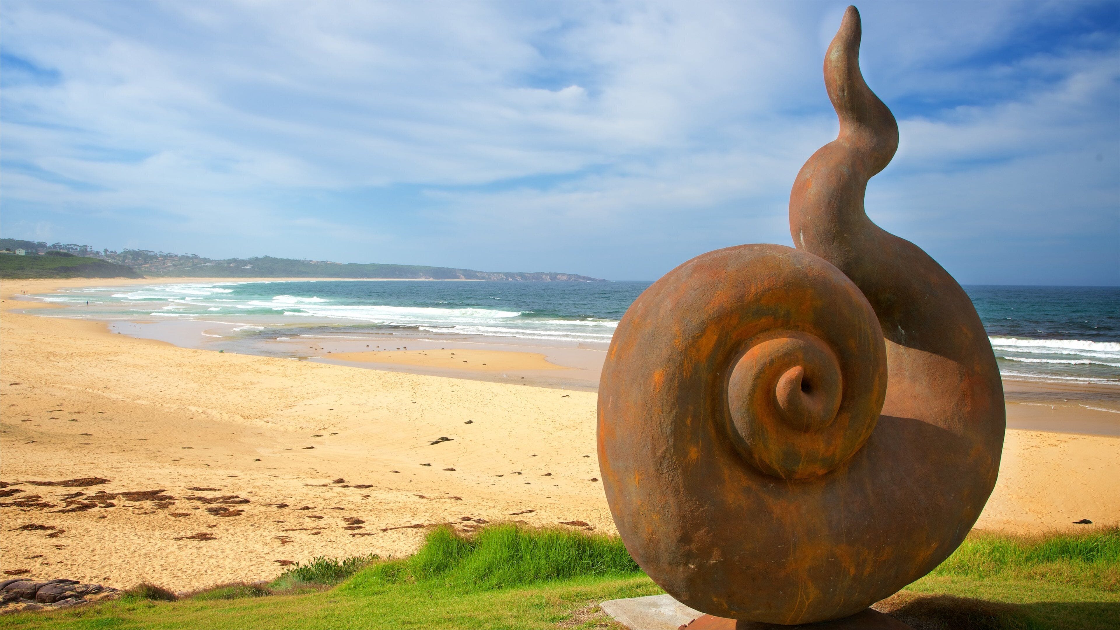 Una gran escultura oxidada de caracol en una playa, con olas de fondo.