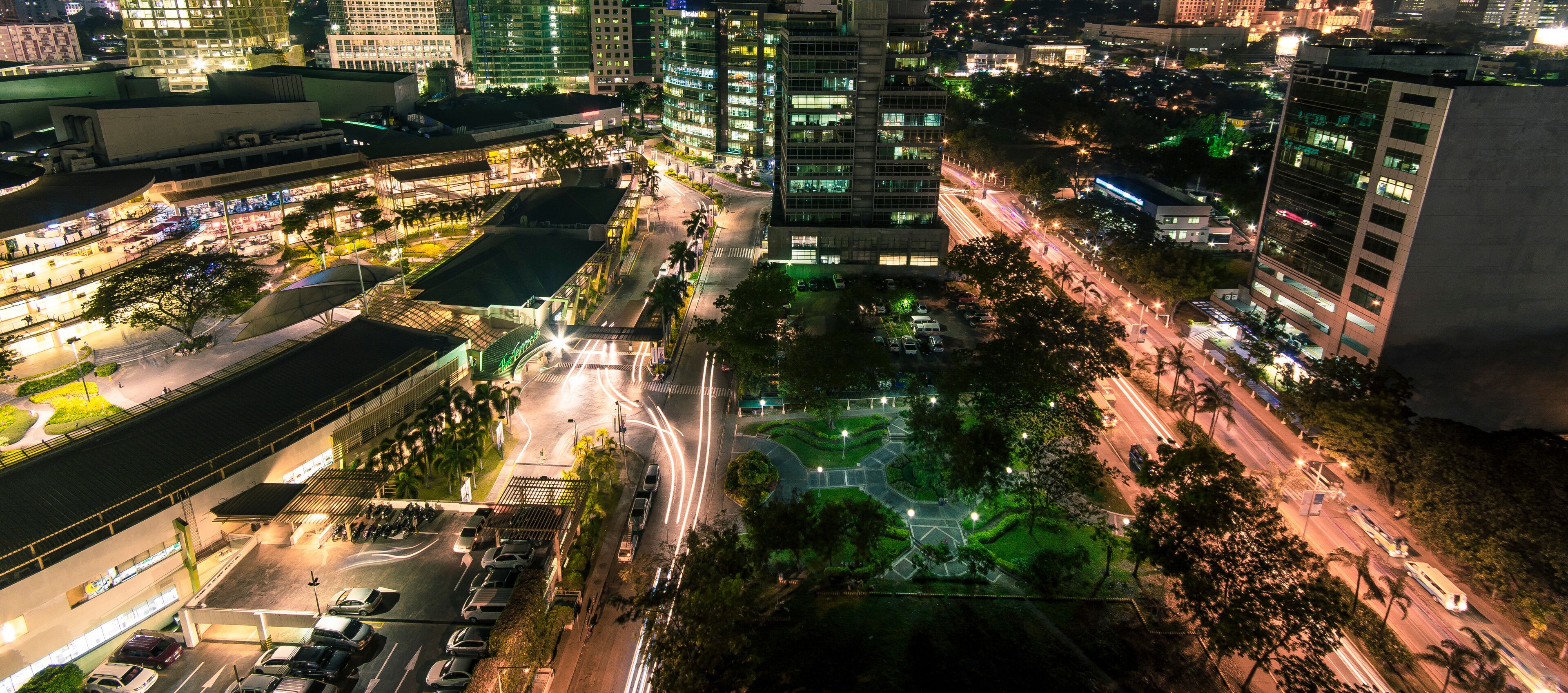 A nighttime cityscape with illuminated buildings, a central roundabout, and surrounding greenery.