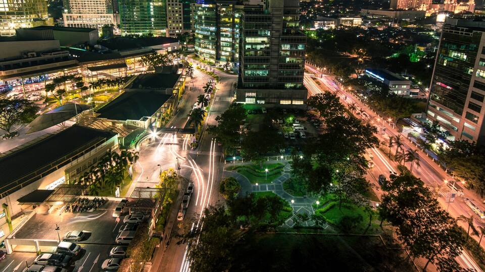 A nighttime cityscape with illuminated buildings, a central roundabout, and surrounding greenery.