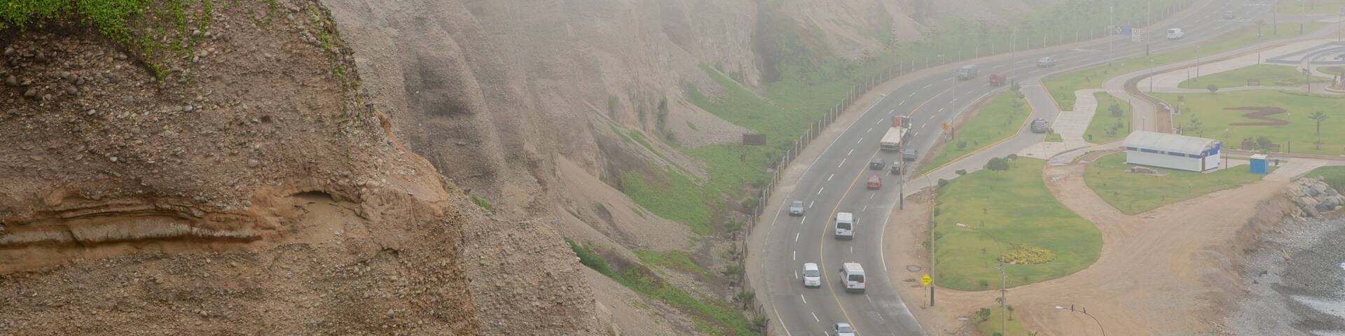 A highway curving through a mountainous landscape with a steep cliff on one side and a grassy area with a building on the other.