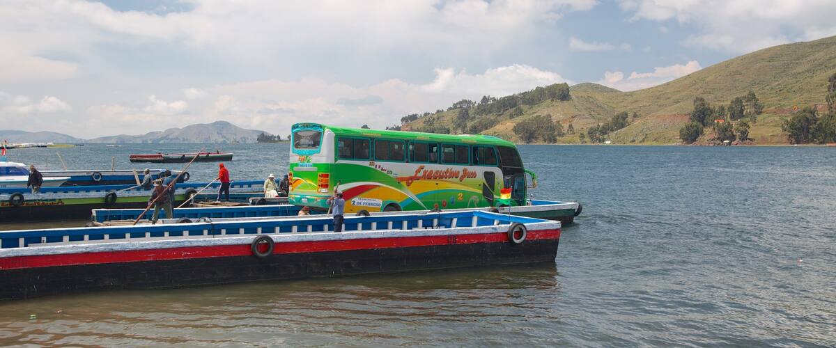 A green and white bus on a lake with a mountainous backdrop.
