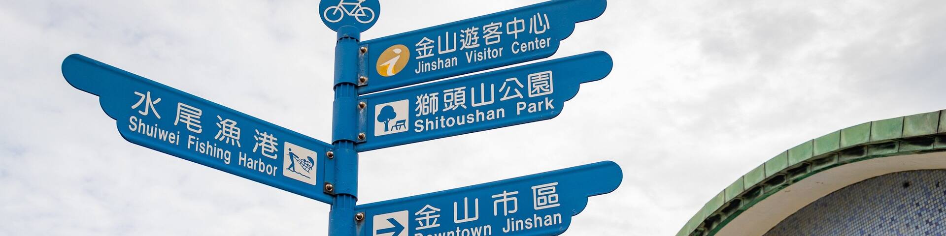 A blue signpost with multiple directions, including Shuei Fishing Harbor, Jinshan Visitor Center, Shitoushan Park, and Downtown Jinshan.