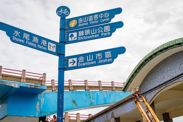 A blue signpost with multiple directions, including Shuei Fishing Harbor, Jinshan Visitor Center, Shitoushan Park, and Downtown Jinshan.