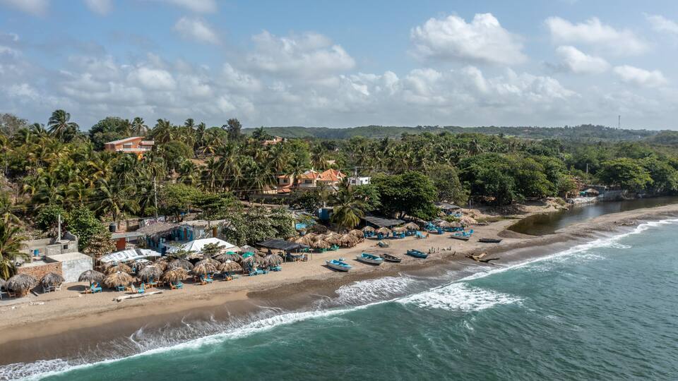 A coastal beach with thatched-roof huts, palm trees, and a clear sky.