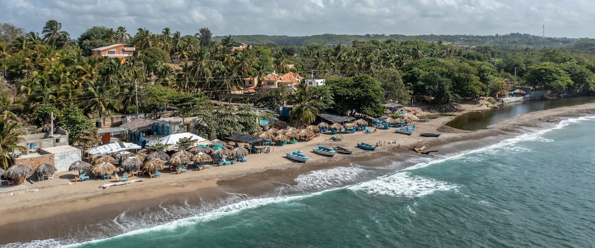 A coastal beach with thatched-roof huts, palm trees, and a clear sky.