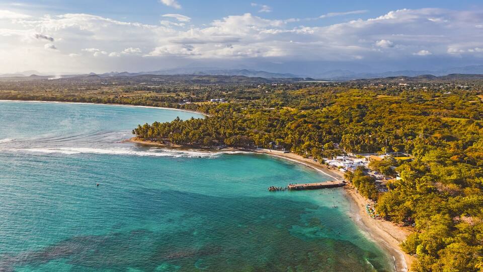 A coastal landscape with a turquoise bay, a pier, and a densely forested area.
