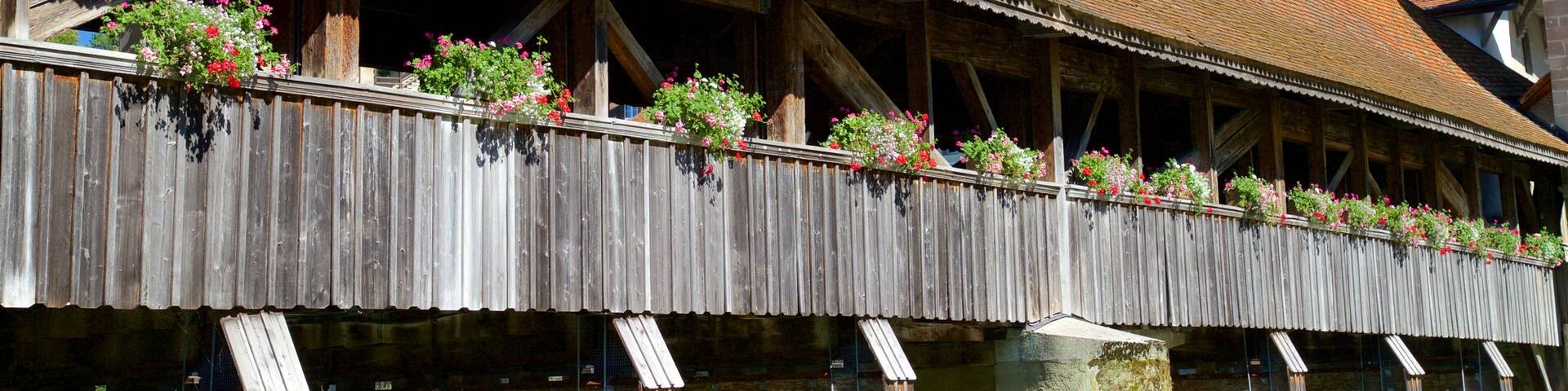 A wooden covered bridge with flower boxes and a tower in the background.