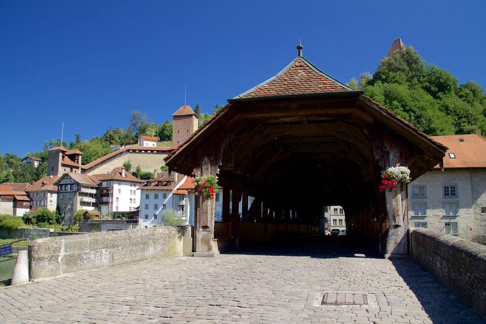 Un puente cubierto histórico con muros de piedra y cestas de flores, rodeado de edificios tradicionales europeos y un río.