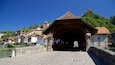 A historic covered bridge with stone walls and flower baskets, surrounded by traditional European buildings and a river.