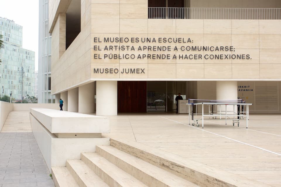 A modern building with a sign that reads "MUSEO JUMEX" and a message about the museum being a school. There are steps leading up to the entrance and a tennis table in the foreground.
