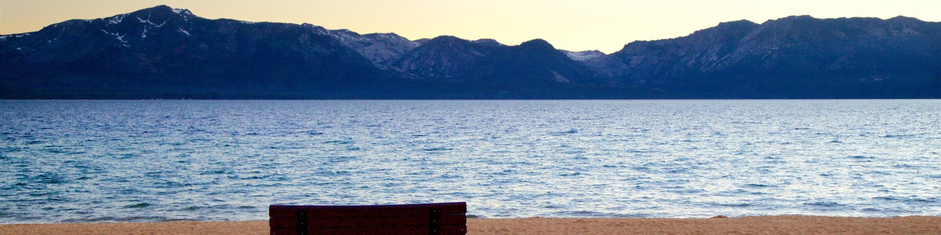 Un banco en una playa arenosa, con un mar tranquilo y montañas al fondo.