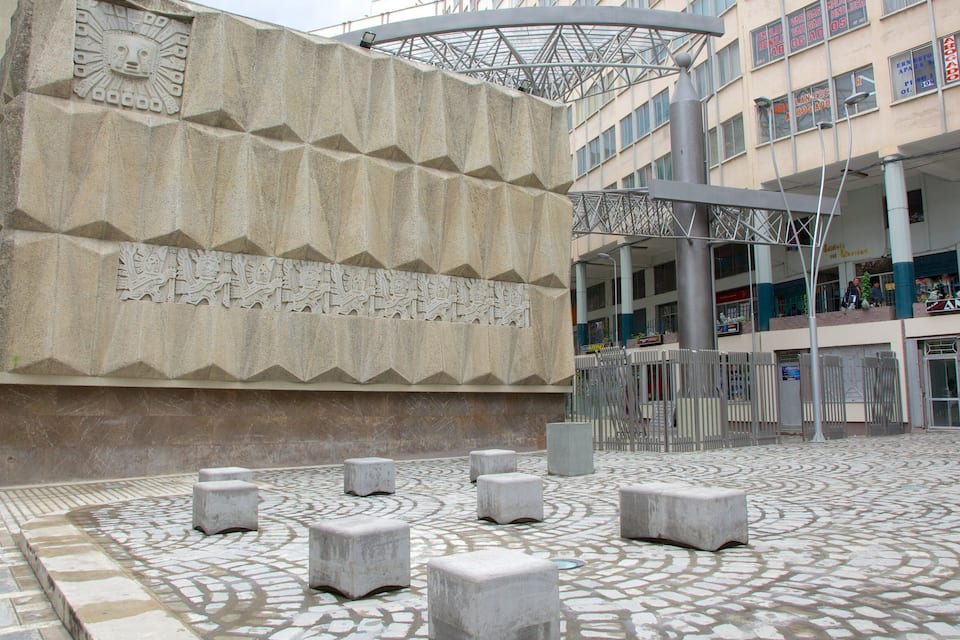 Uma praça com um grande monumento de pedra, detalhado com esculturas complexas e um piso de azulejos estampados.