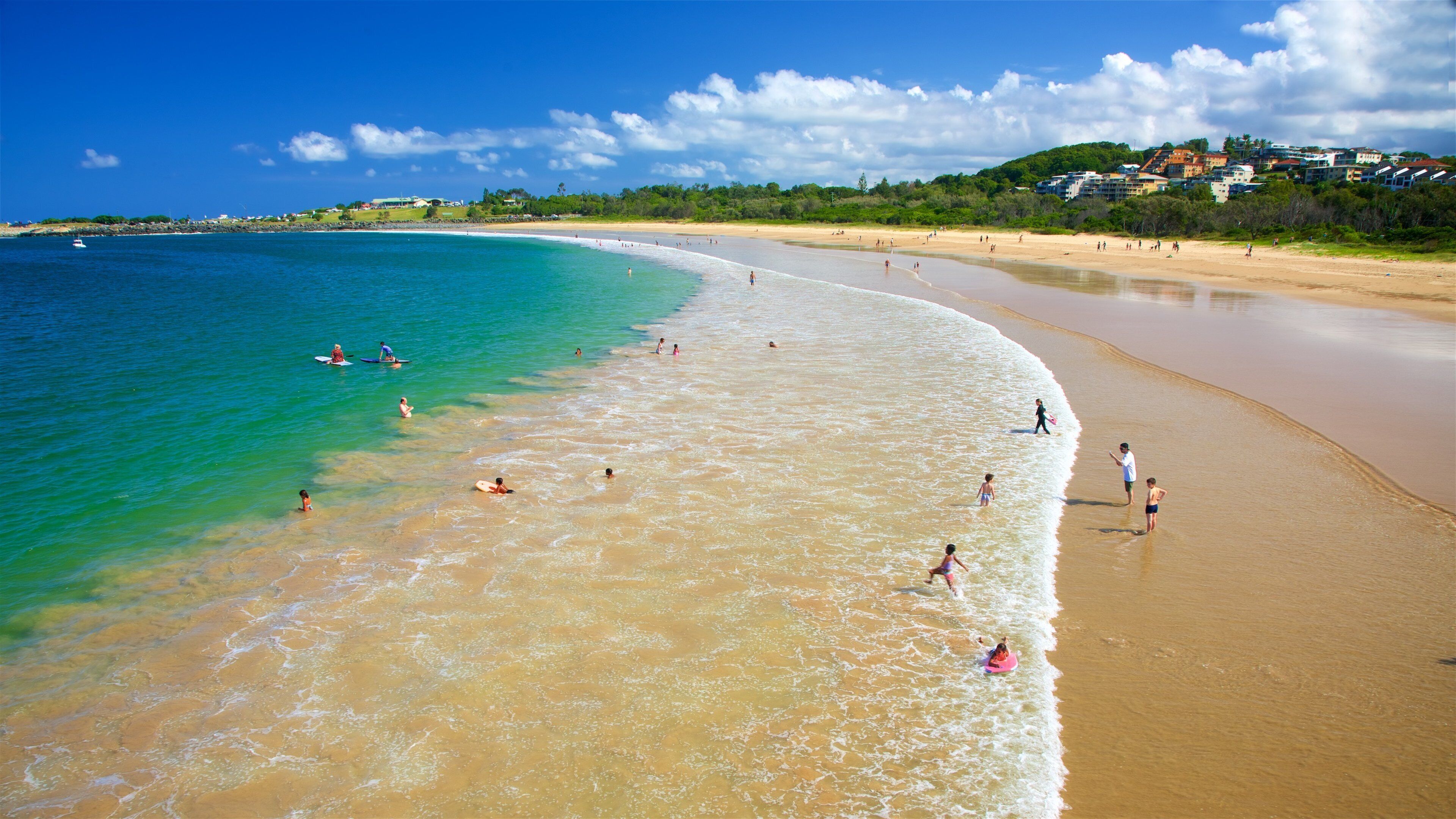 A beach with clear turquoise water, a sandy shore, and buildings in the background.