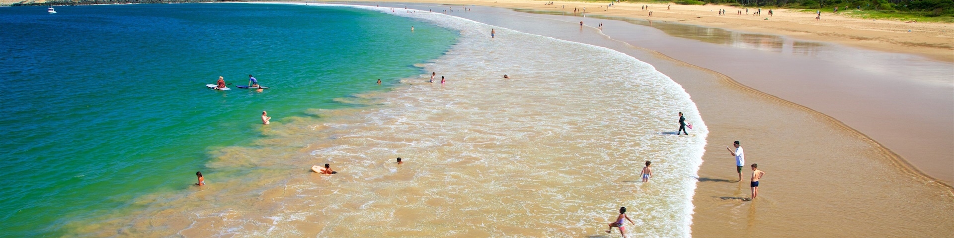 A beach with clear turquoise water, a sandy shore, and buildings in the background.
