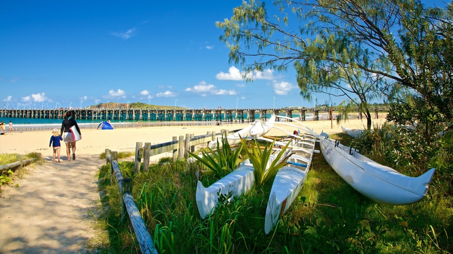 Una playa con un muelle de madera, canoas blancas y una mujer caminando con un niño.