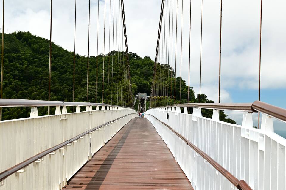 Un pont de fusta per a vianants amb baranes i cables blancs, que condueix a una construcció allunyada.