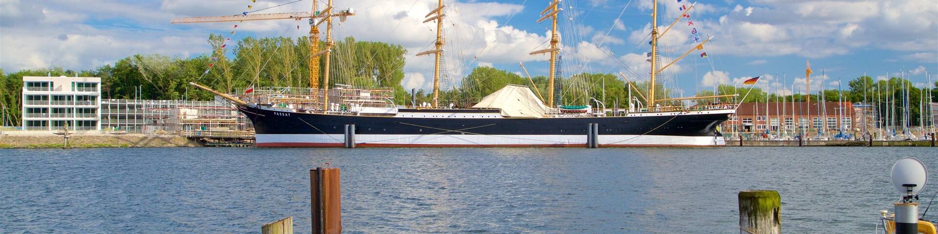 A large sailing ship docked at a harbor with a building and trees in the background.