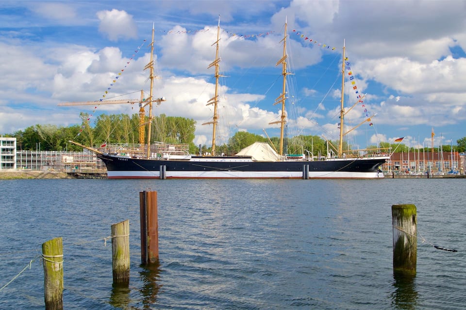 A large sailing ship docked at a harbor with a building and trees in the background.