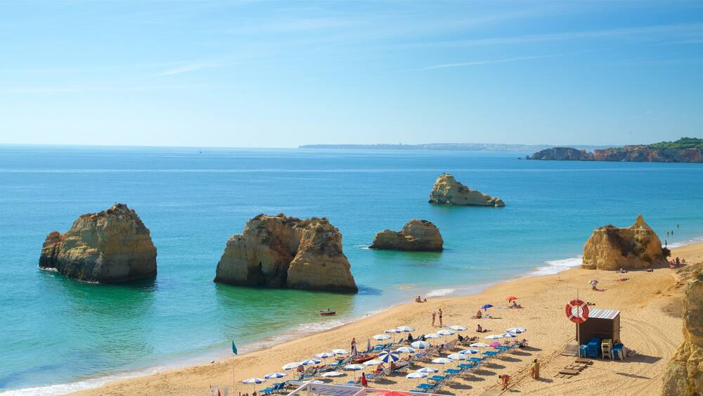 A beach with large rock formations, clear blue water, and a sandy shore with beach umbrellas and chairs.