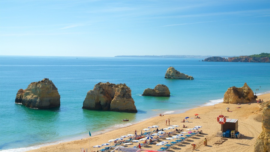 Une plage caractérisée par de grandes formations rocheuses, une eau d’un bleu limpide et un rivage sablonneux agrémenté de parasols et de chaises de plage.