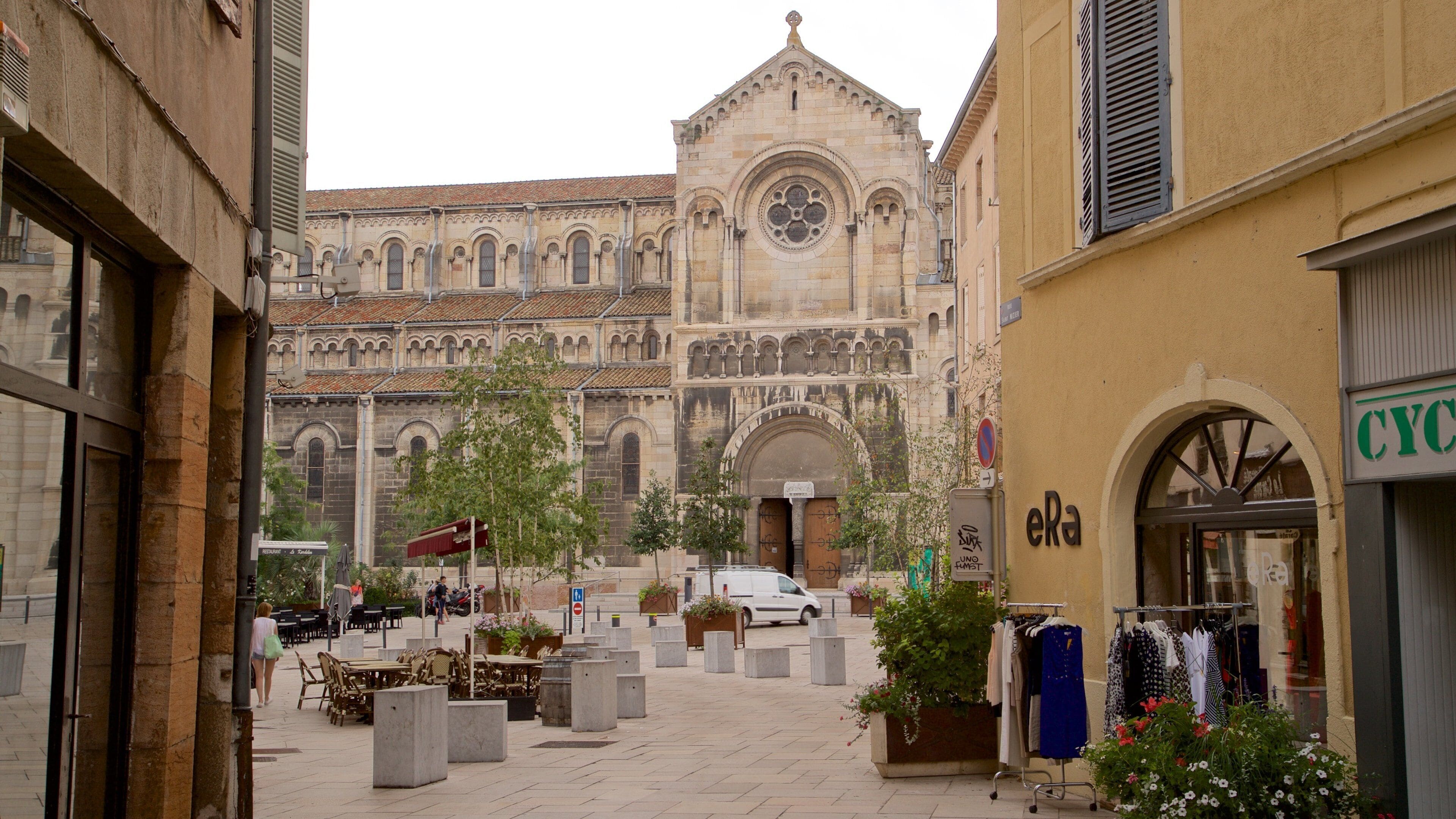 Un patio con un edificio histórico al fondo, mobiliario exterior y una tienda.