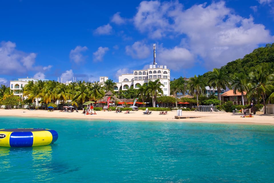 A beach with clear turquoise water, palm trees, and a white building with arched windows in the background.