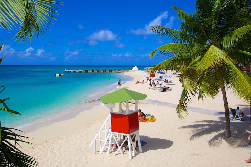 Une plage avec une eau turquoise et limpide, du sable blanc, des palmiers et une tour de surveillance pour les sauveteurs.