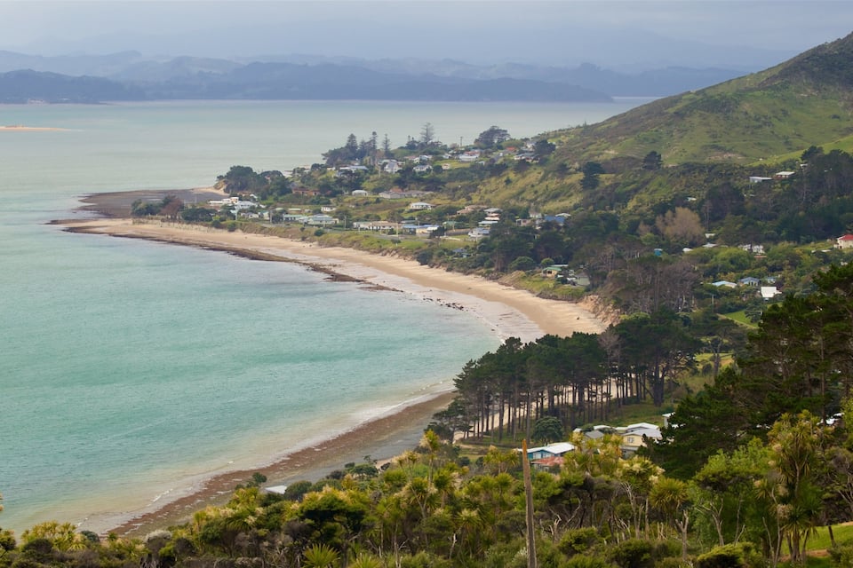 A coastal landscape with a sandy beach, a calm sea, and a backdrop of hills and scattered houses.