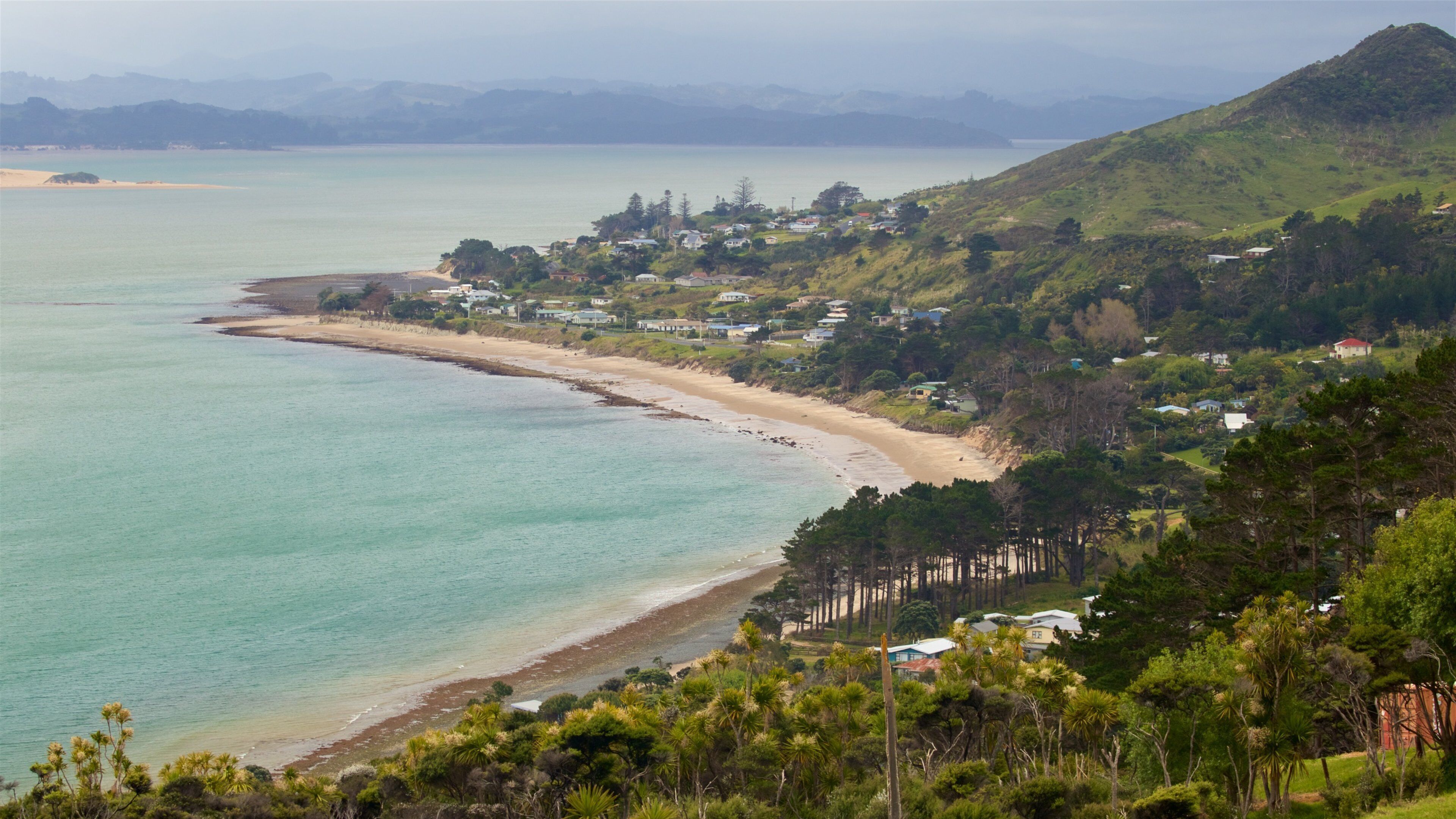 A coastal landscape with a sandy beach, a calm sea, and a backdrop of hills and scattered houses.