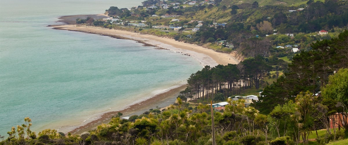 A coastal landscape with a sandy beach, a calm sea, and a backdrop of hills and scattered houses.