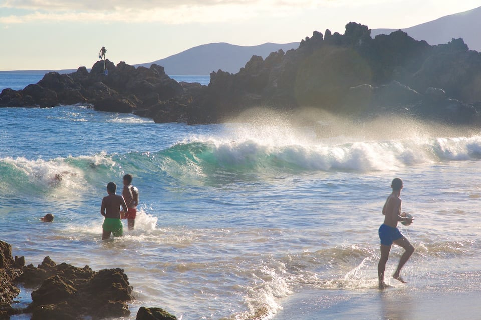 Una playa con olas, afloramientos rocosos y montañas al fondo.