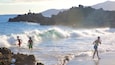 A beach with waves, rocky outcrops, and mountains in the background.