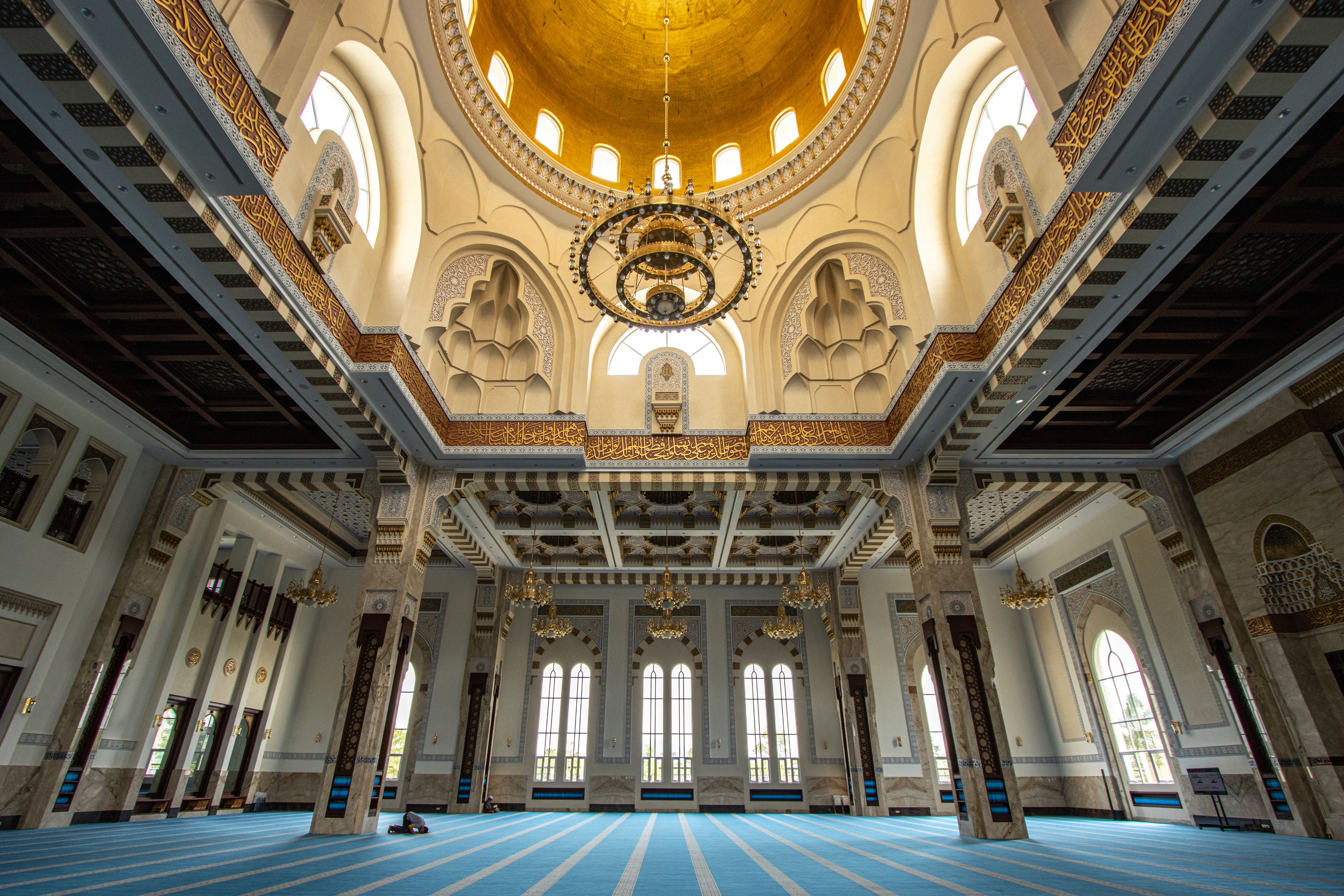 An interior of a mosque with blue carpet, golden dome, and intricate designs.