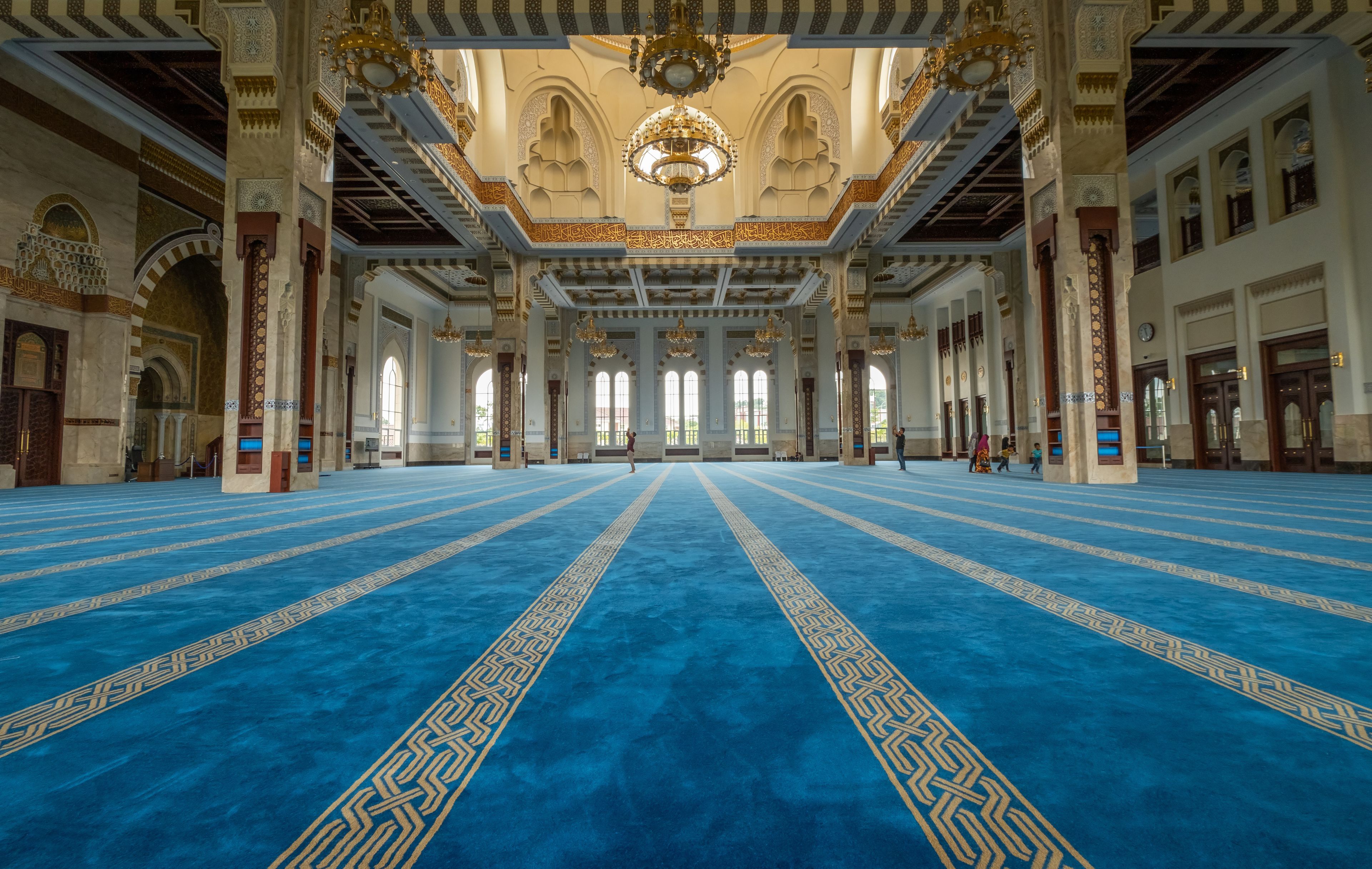 A large hall with blue carpet and intricate golden patterns, high ceilings with ornate chandeliers, and tall columns.