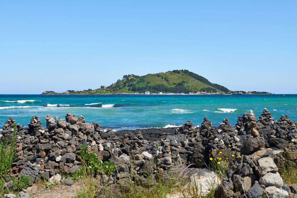 Un rivage rocheux où les vagues s’écrasent sur la plage, une petite colline avec des bâtiments visibles au loin, et un ciel bleu dégagé.