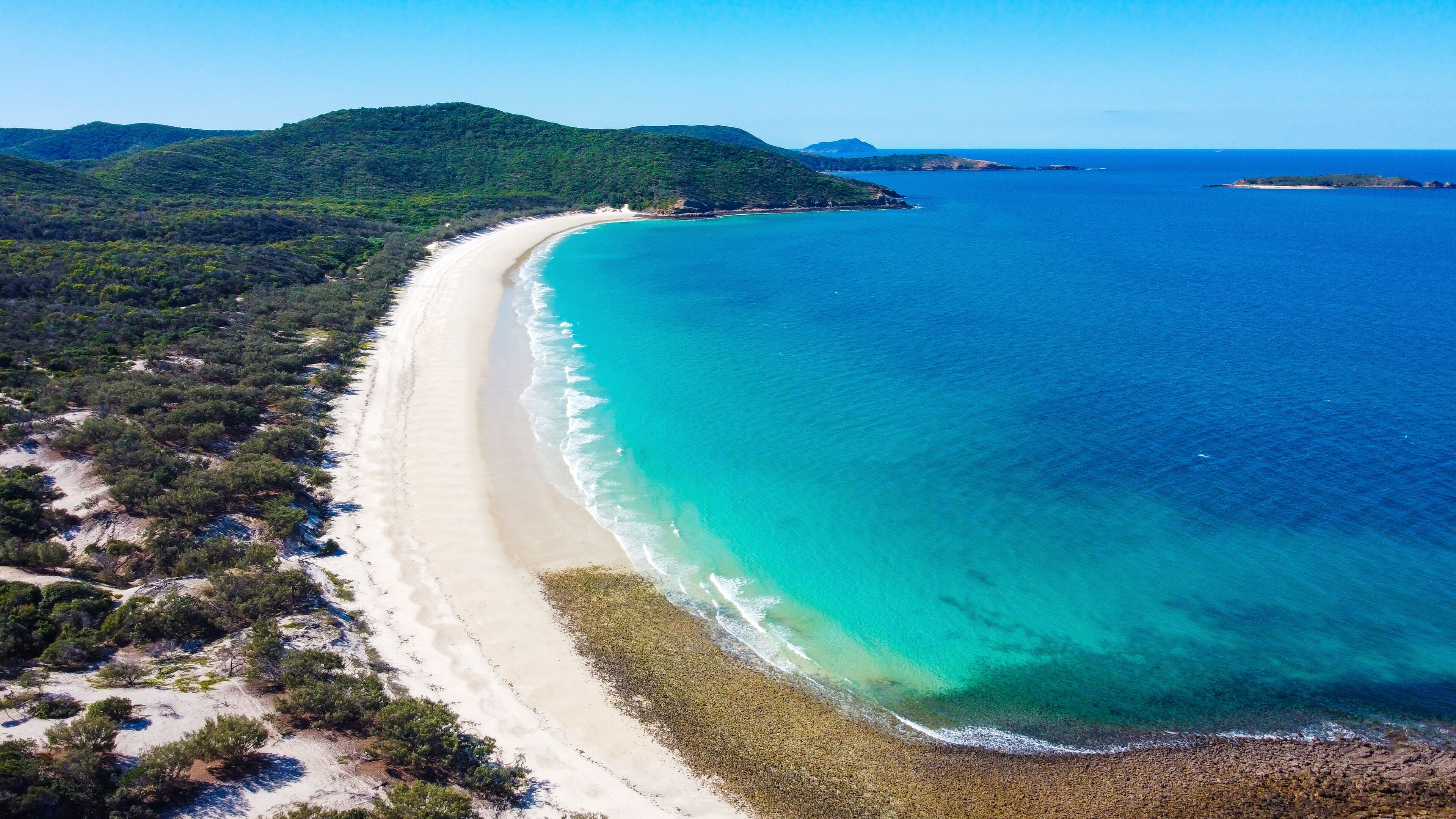 A coastal landscape with a long sandy beach, clear turquoise waters, and green hills in the background.