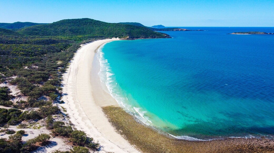 A coastal landscape with a long sandy beach, clear turquoise waters, and green hills in the background.