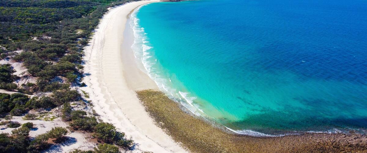 A coastal landscape with a long sandy beach, clear turquoise waters, and green hills in the background.