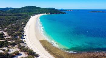 Eine Küstenlandschaft mit einem langen Sandstrand, klarem türkisblauem Wasser und grünen Hügeln im Hintergrund.