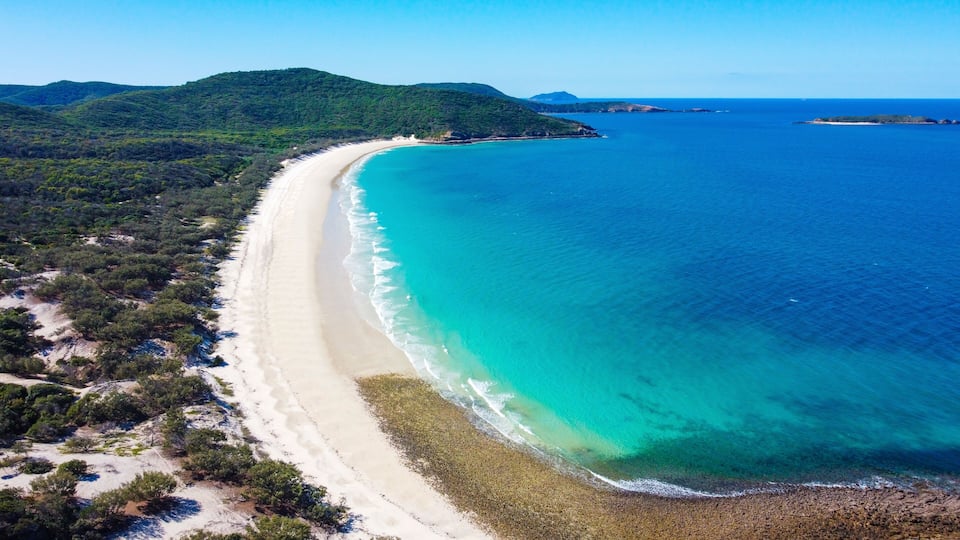 A coastal landscape with a long sandy beach, clear turquoise waters, and green hills in the background.