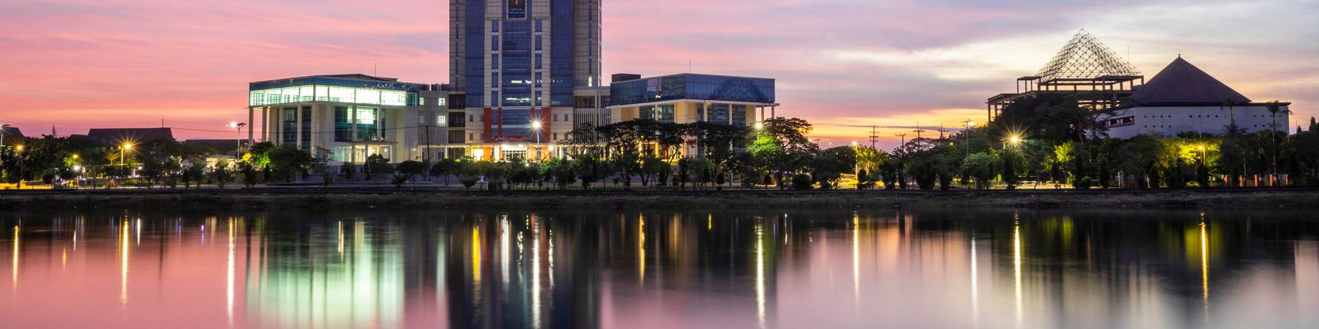 A cityscape at dusk with a river reflecting buildings, a modern skyscraper, and a traditional structure with a pyramid-like roof.