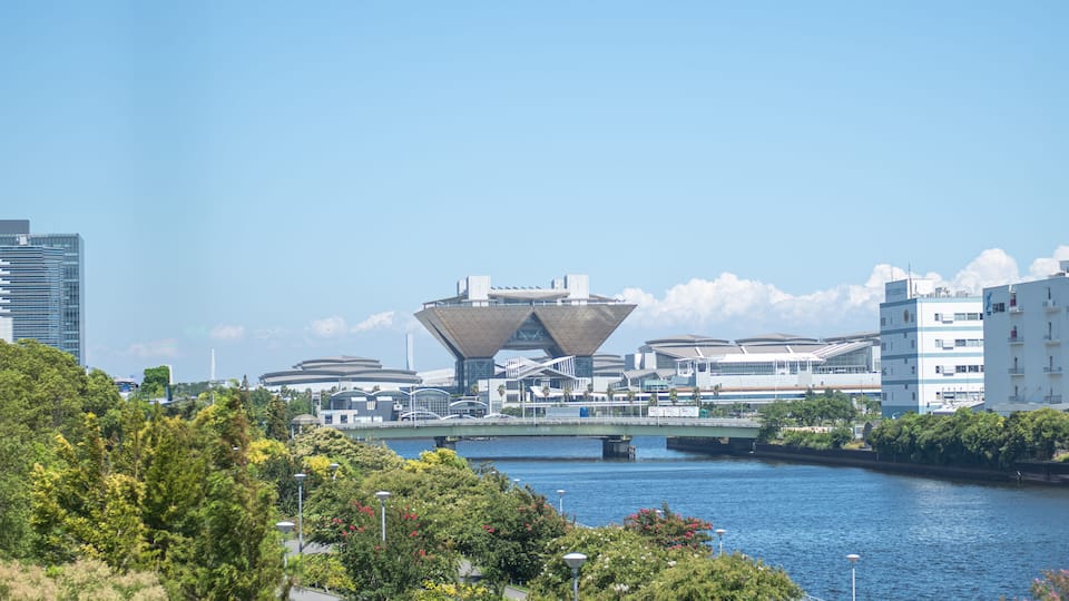 Uma estrutura arquitetônica moderna junto a um rio, cercada por vegetação e edifícios urbanos.